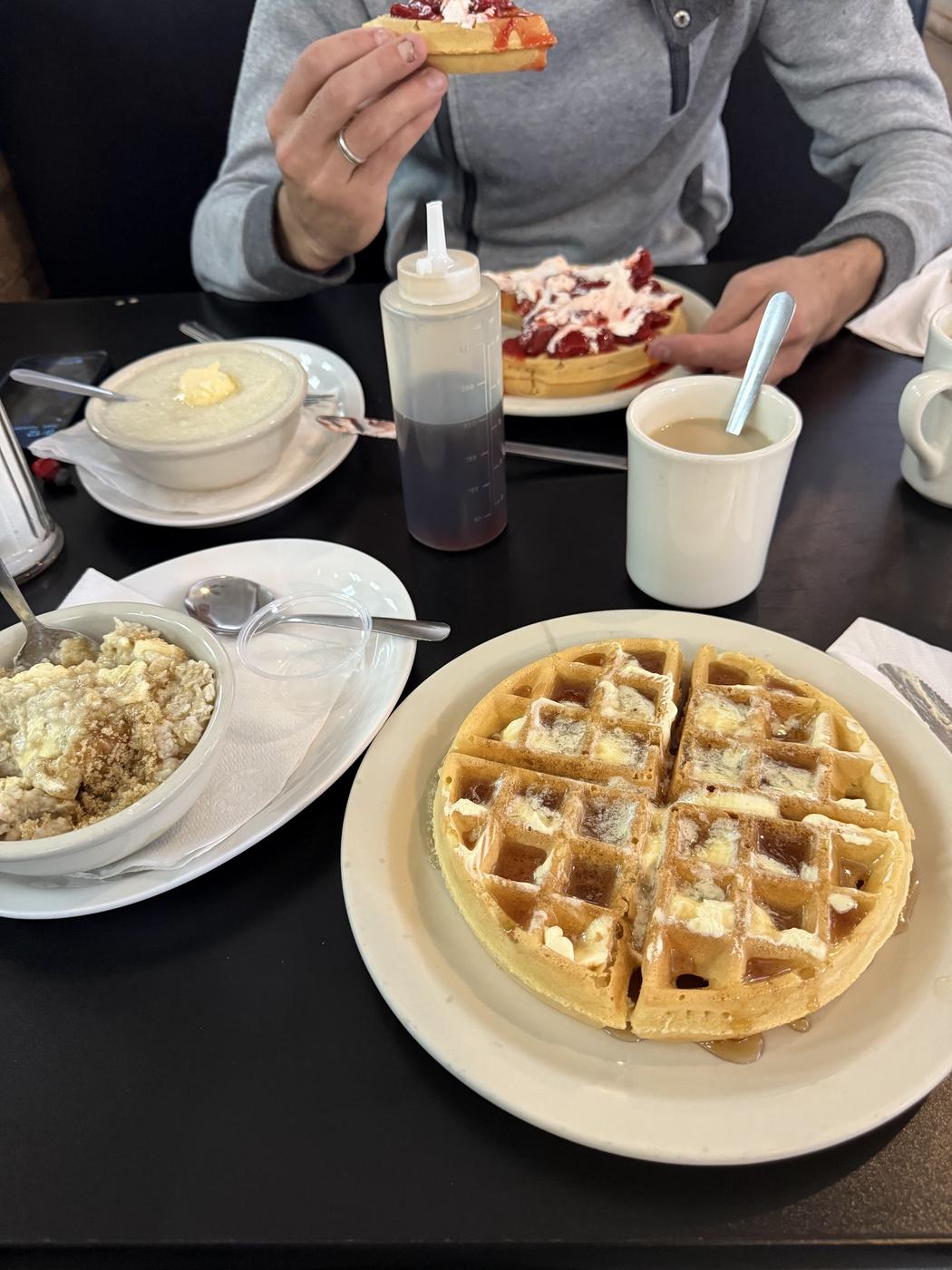 A waffle with butter and syrup on a plate, a bowl of grits and a bowl of oatmeal with brown sugar